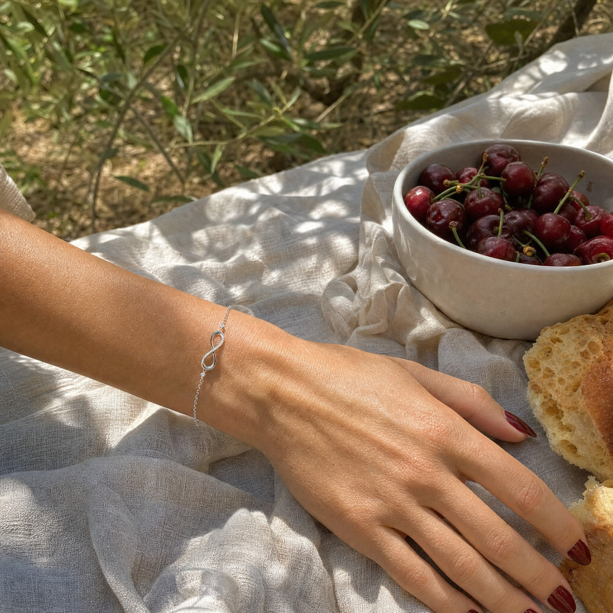 Her hand resting on a linen blanket at an olive-grove picnic beside cherries and bread, the infinity bracelet at her wrist.