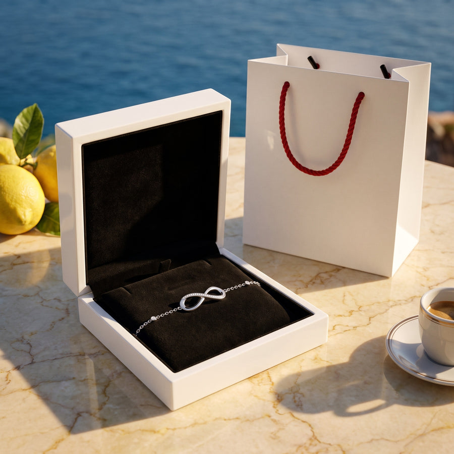 The open white box (black velvet inside) and the white bag with red rope handles on a sunlit terrace table, warm shadows.