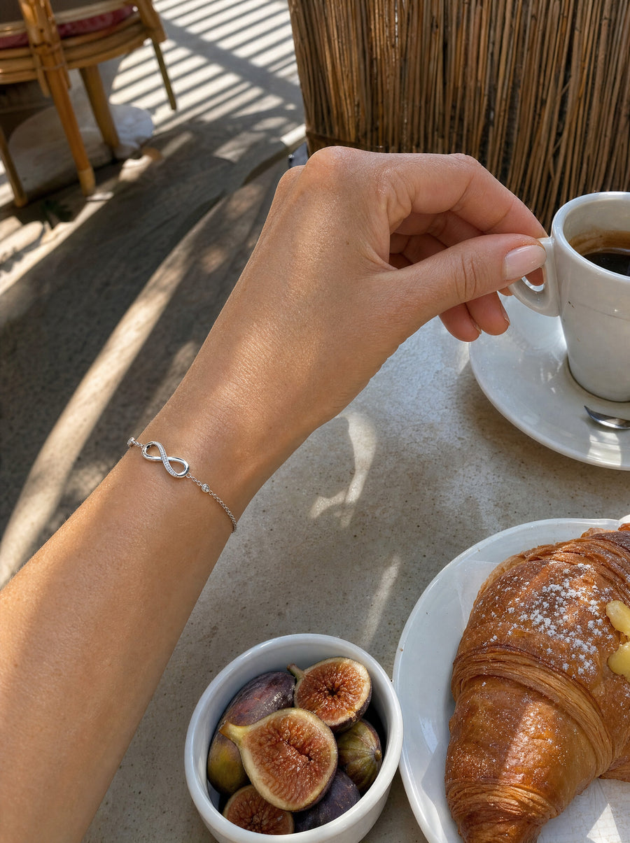 Her hand lifting an espresso cup at a sunny cafe table with a croissant and fresh figs, the infinity bracelet at her wrist.