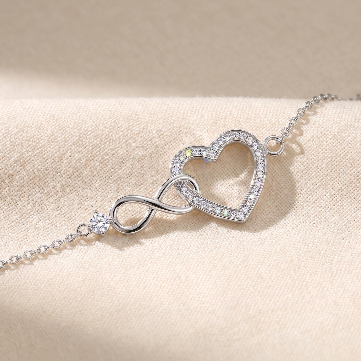 The Mossante Infinity Heart Bracelet laid on a warm cream linen cloth: a fine cable chain in silver curving in from both edges to one ornament at the center, a round moissanite in prongs at the tip of a polished infinity, the infinity linked through an open heart lined with one row of small stones.