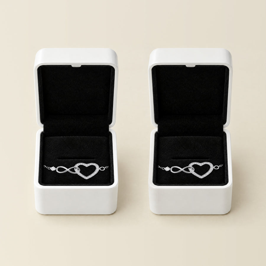 Two silver infinity heart bracelets side by side on a cream ground, each in its own open white box with a black velvet interior.