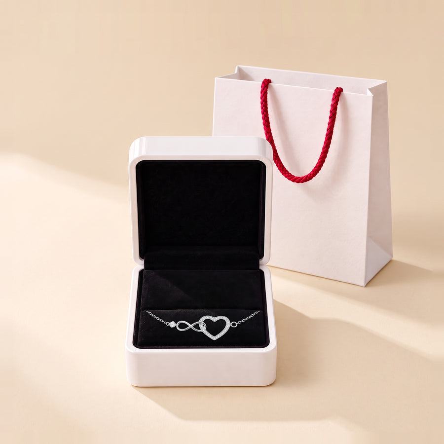 The infinity heart bracelet in its white box with the lid open, the heart and the infinity on the black velvet pad, a white paper bag with red rope handles beside it on a cream ground.