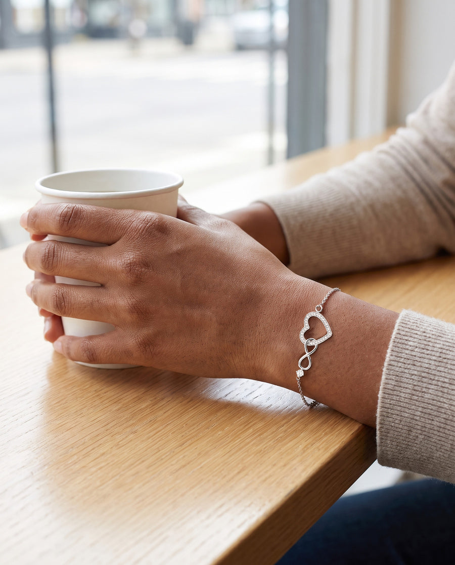 A warm-brown-skinned woman's hand round a plain white cup on a pale wood cafe table by a window in morning light, an oat knit sleeve at the wrist, the silver infinity heart bracelet on top of her wrist.