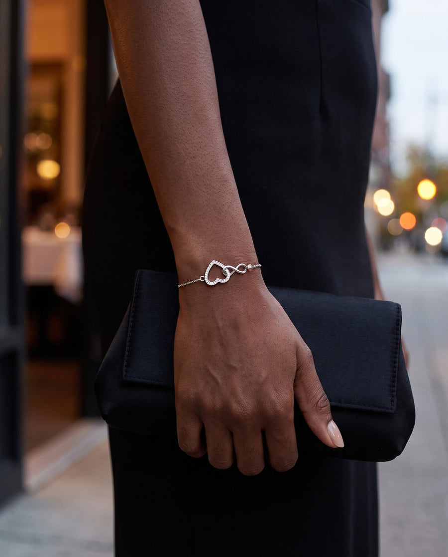 A Black woman's hand holding a black satin clutch against a black dress, warm restaurant lights blurred behind at dusk, the open heart and the infinity catching the light at her wrist.