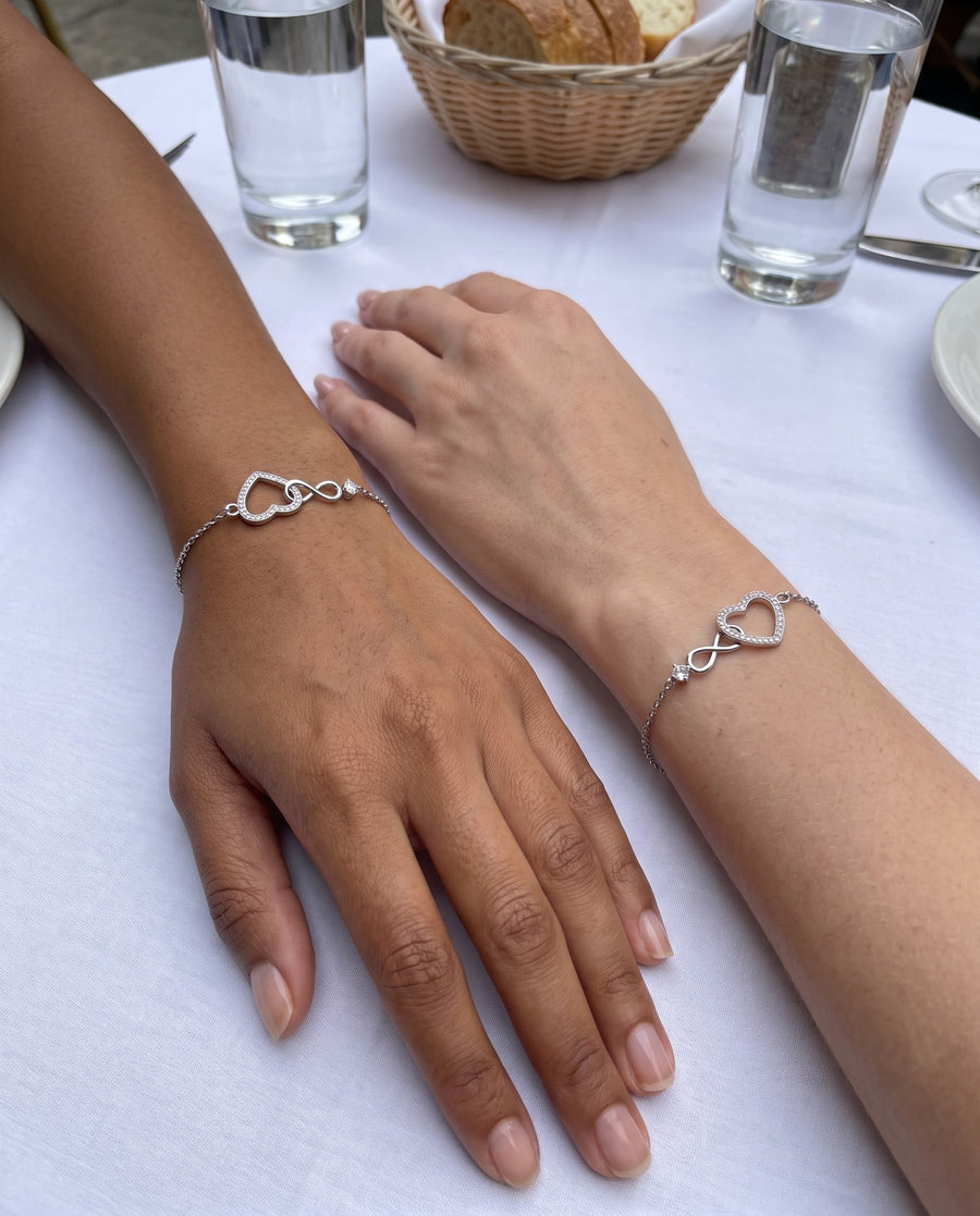 Two women's wrists side by side on a white linen brunch table in daylight, one olive-skinned and one light-skinned, each wearing the silver infinity heart bracelet, glasses of water behind.