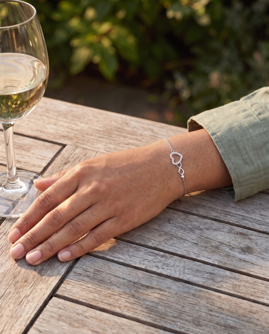 A Southeast Asian woman's wrist with medium tan skin resting beside a glass of white wine on a weathered teak garden table in late afternoon sun, a sage linen sleeve, the open heart bright on top of her wrist.