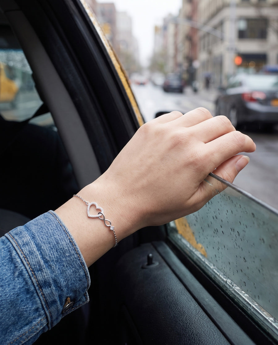 A light-skinned woman's hand resting on the open window frame of a taxi, a city street blurred behind in overcast daylight, a denim jacket cuff, the silver bracelet on top of her wrist.