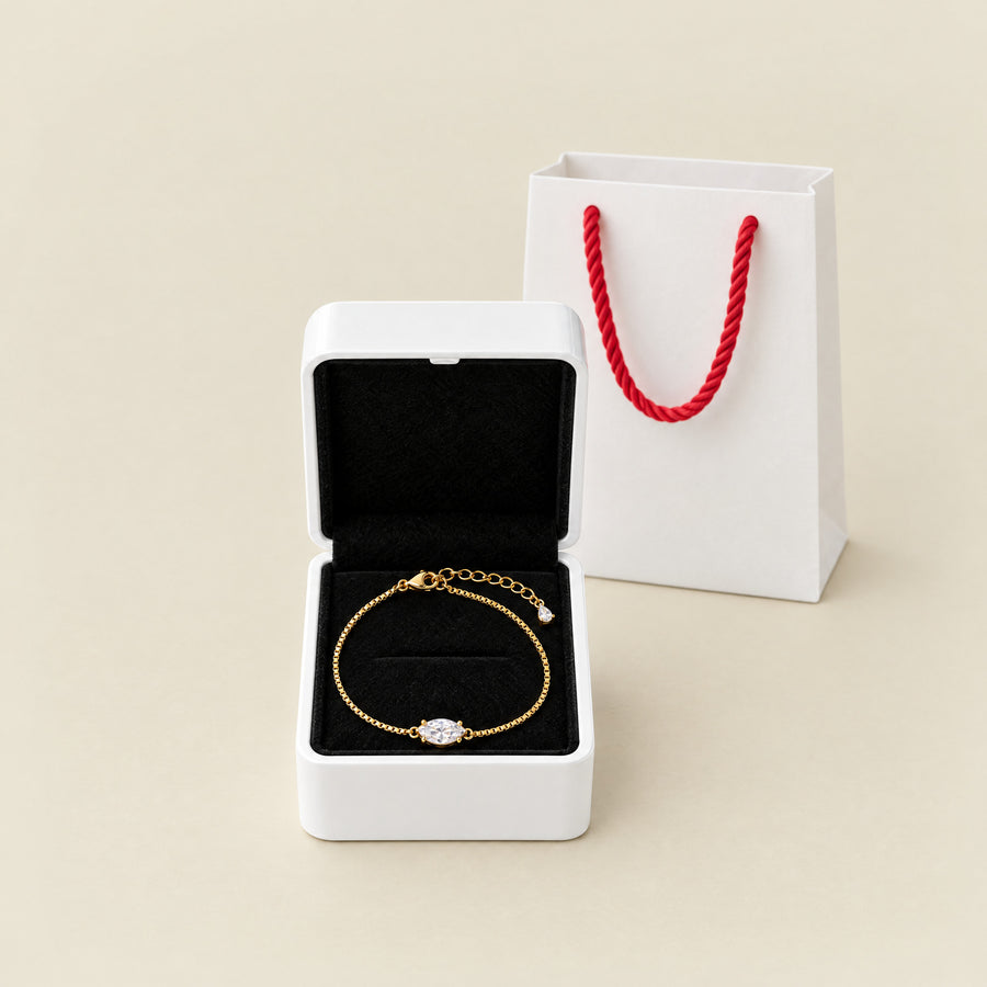 The marquise bracelet in its white box with the lid open, the black velvet interior holding the chain, a white paper bag with red rope handles beside it on a cream ground.
