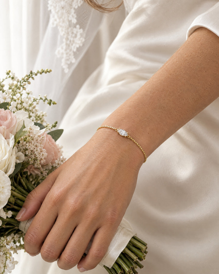 A bride's hand holding the stems of a small white bouquet against ivory silk, the gold plated marquise bracelet at the wrist, the stone catching daylight.