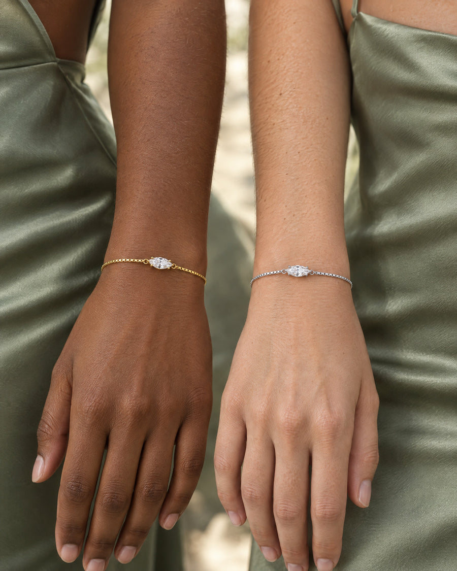 Two women's wrists in sage green satin side by side, each wearing the marquise bracelet, one gold plated and one silver, the stones lined up across the frame.