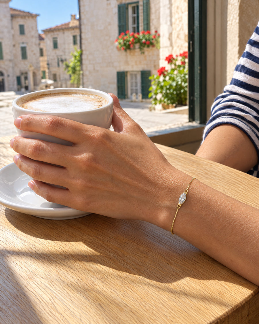 A woman's hand round a cappuccino cup at an open cafe window on a sunny old-town square, the gold plated marquise bracelet on top of her wrist.
