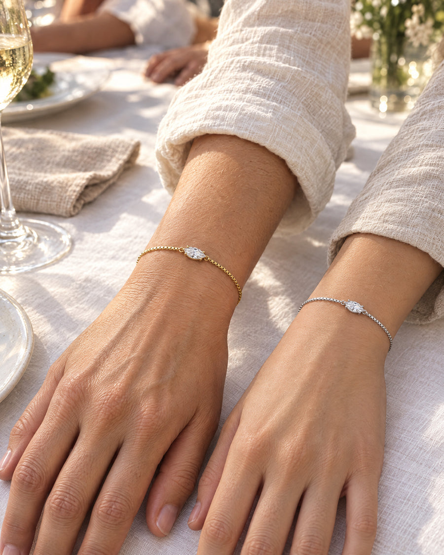 A mother's and a daughter's hands resting side by side on white linen at a garden lunch, one wrist wearing the gold plated bracelet and the other the silver.