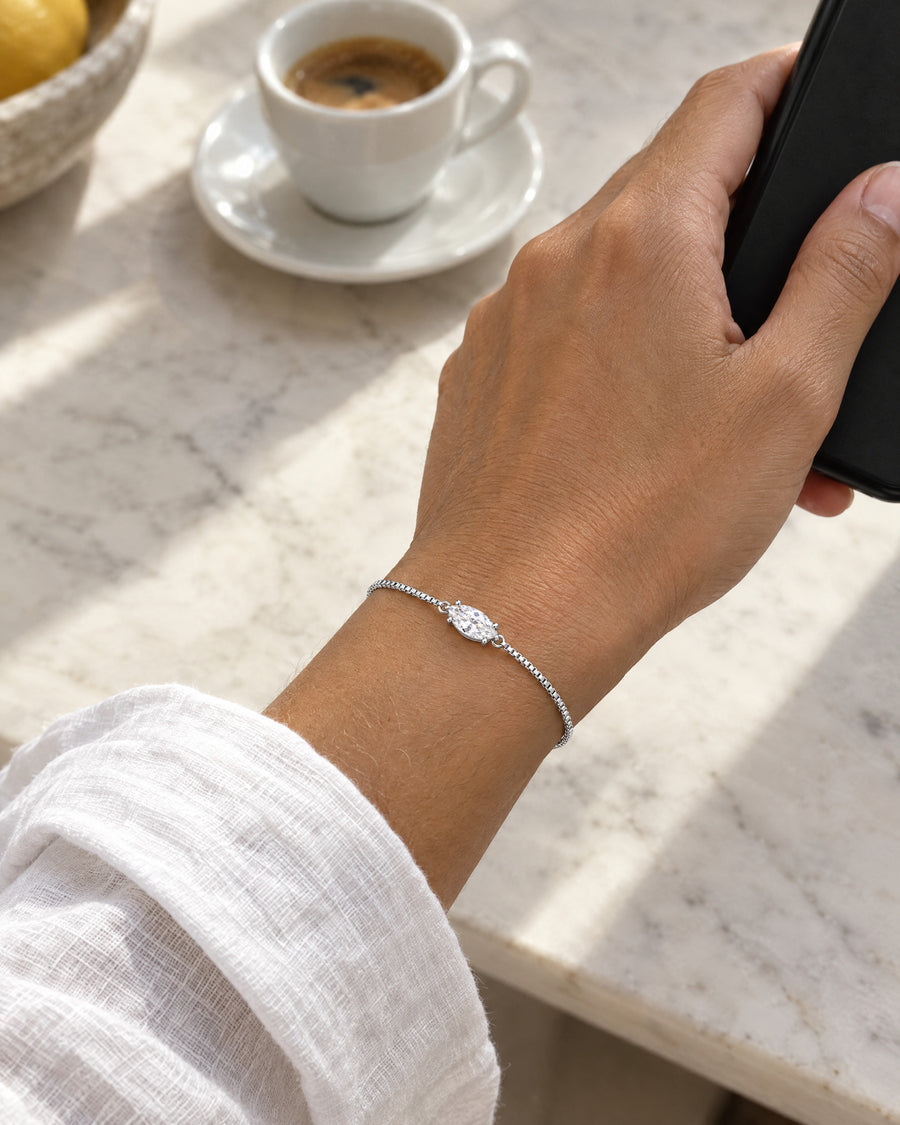 A South Asian woman's hand holding a black phone over a marble counter, an espresso behind, the silver marquise bracelet lying flat at the wrist.