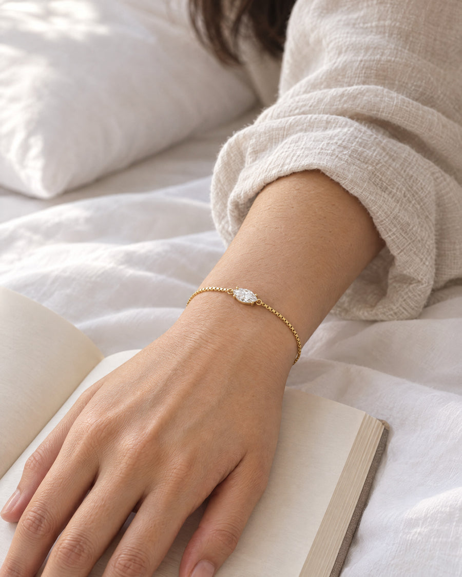 An East Asian woman's hand resting on the open page of a book on white bed linen in morning light, the bracelet loose at the wrist.