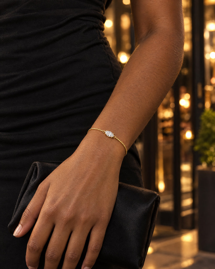 A Black woman's hand holding a black satin clutch against a black dress, restaurant lights behind, the single marquise stone catching the light at the wrist.