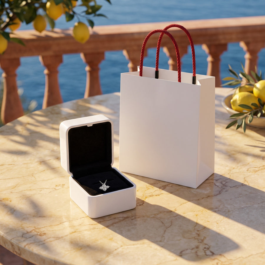 The open white box (black velvet inside) and the white bag with red rope handles on a sunlit terrace table, warm shadows.