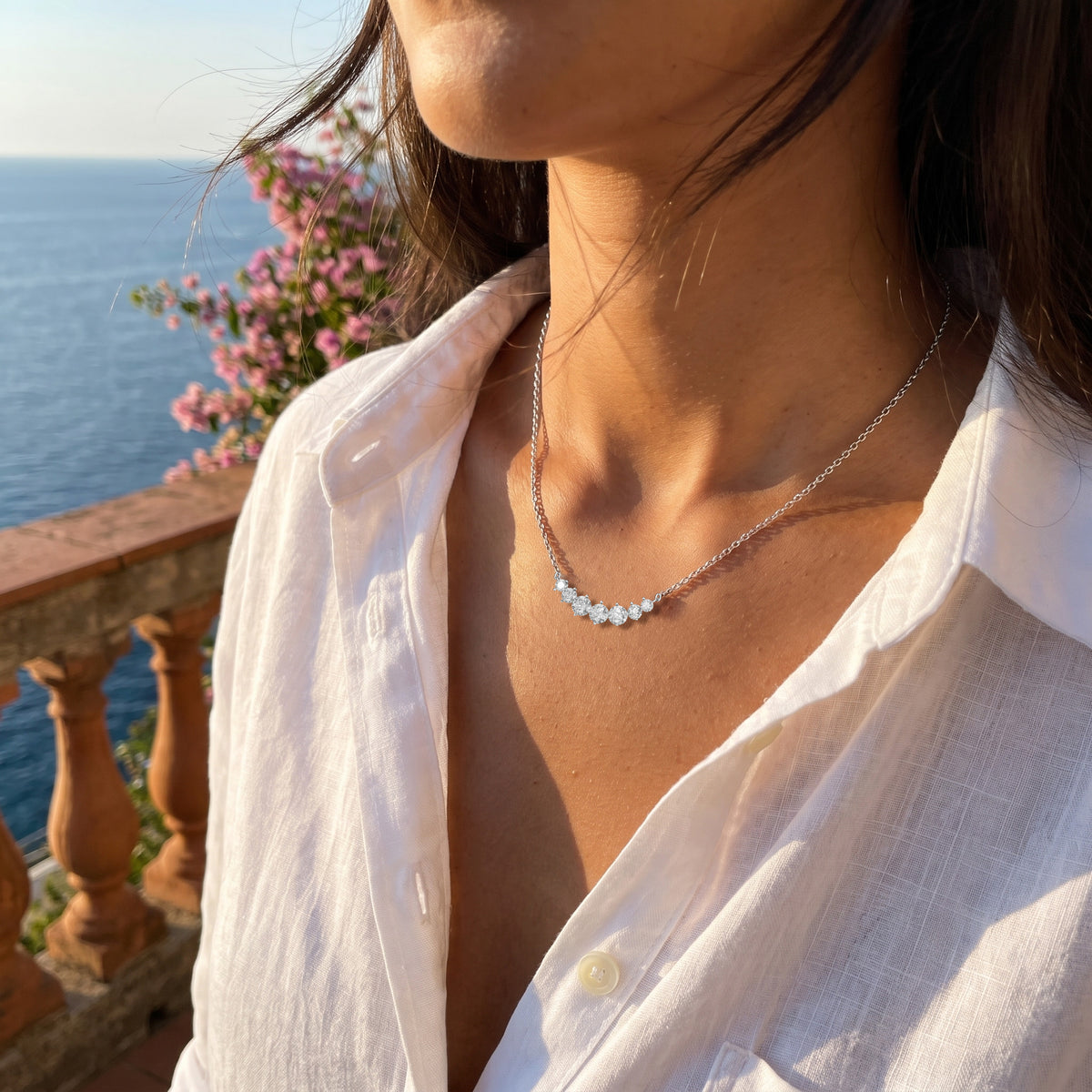 A sunlit terrace above the sea at golden hour, an open white linen shirt, wind in her hair, cropped at the chin.