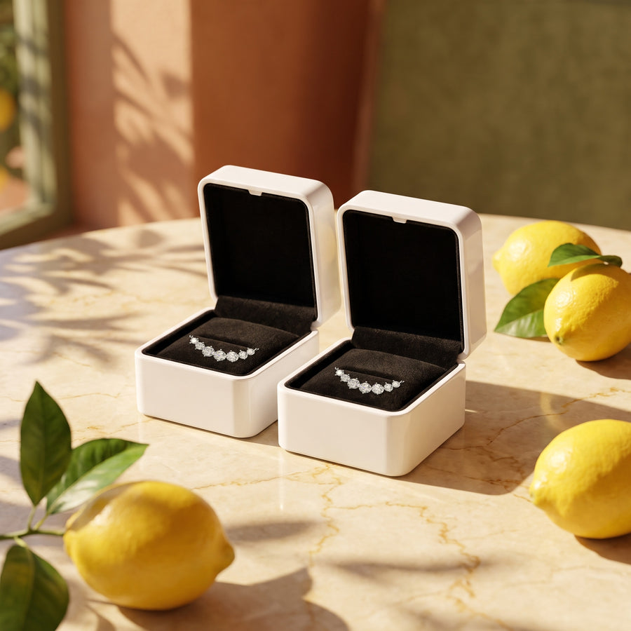 Two open white boxes (black velvet inside) on a sunlit marble table with lemons and leaf shadows.