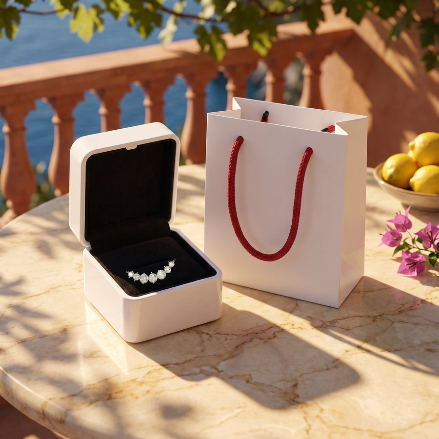 The open white box (black velvet inside) and the white bag with red rope handles on a sunlit terrace table, warm shadows.