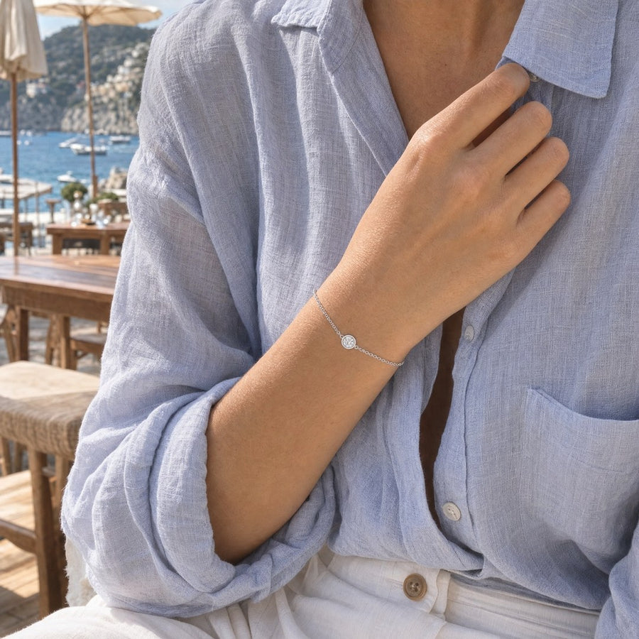 A hand at the collar of a blue linen shirt on a seaside terrace, the silver round-cut bracelet at the wrist.