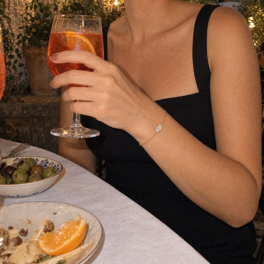 A hand holding an orange spritz at an evening terrace table, the gold plated marquise-cut bracelet at the wrist.