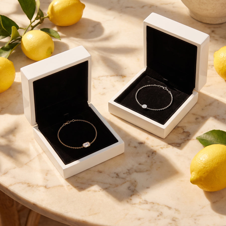 Two open white boxes (black velvet inside) on a sunlit marble table with lemons and leaf shadows.