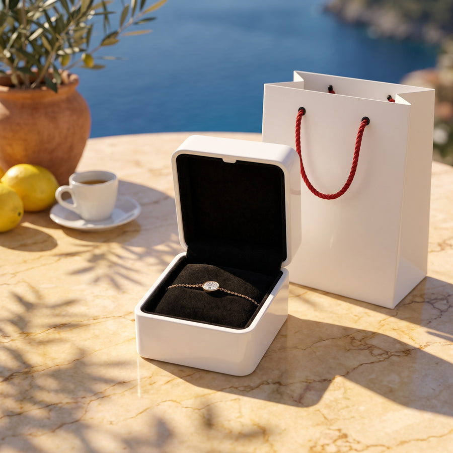 The open white box (black velvet inside) and the white bag with red rope handles on a sunlit terrace table, warm shadows.