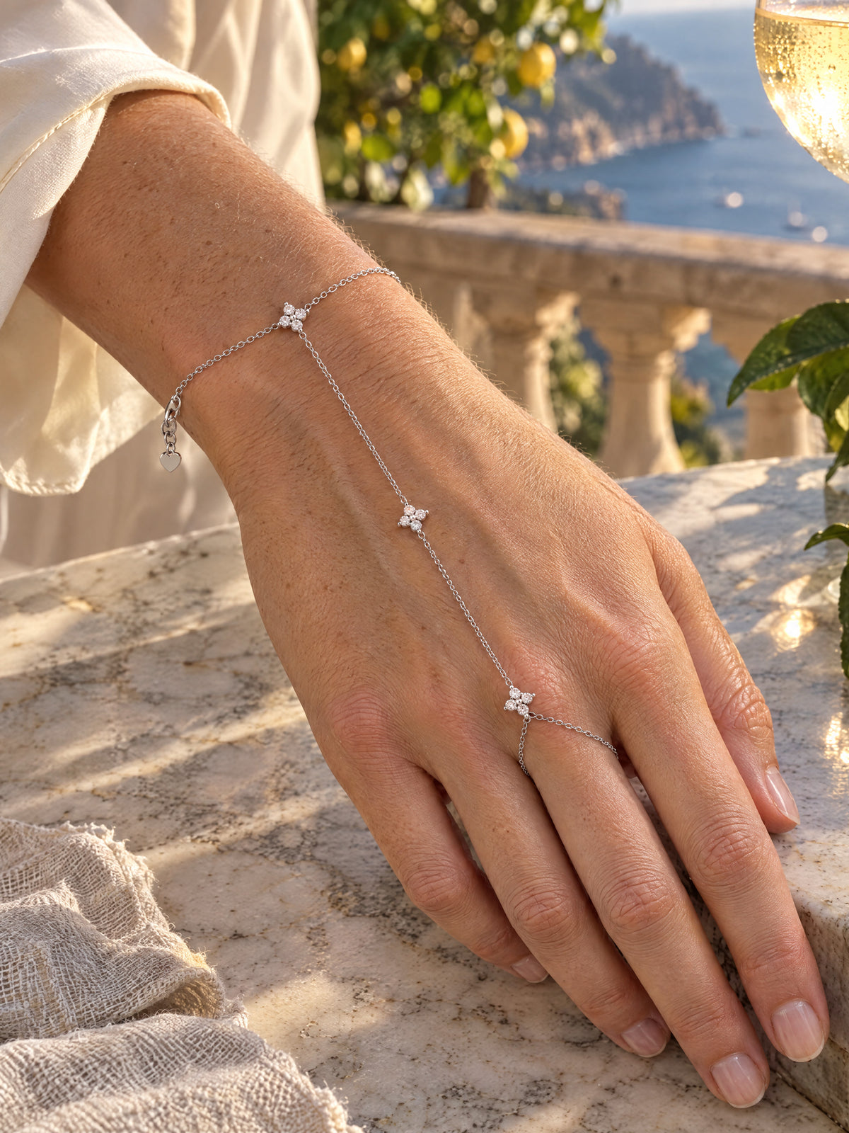 The hand chain worn on a marble table above the Amalfi coast, a lemon tree and a glass of wine behind, the three stars in midday light.