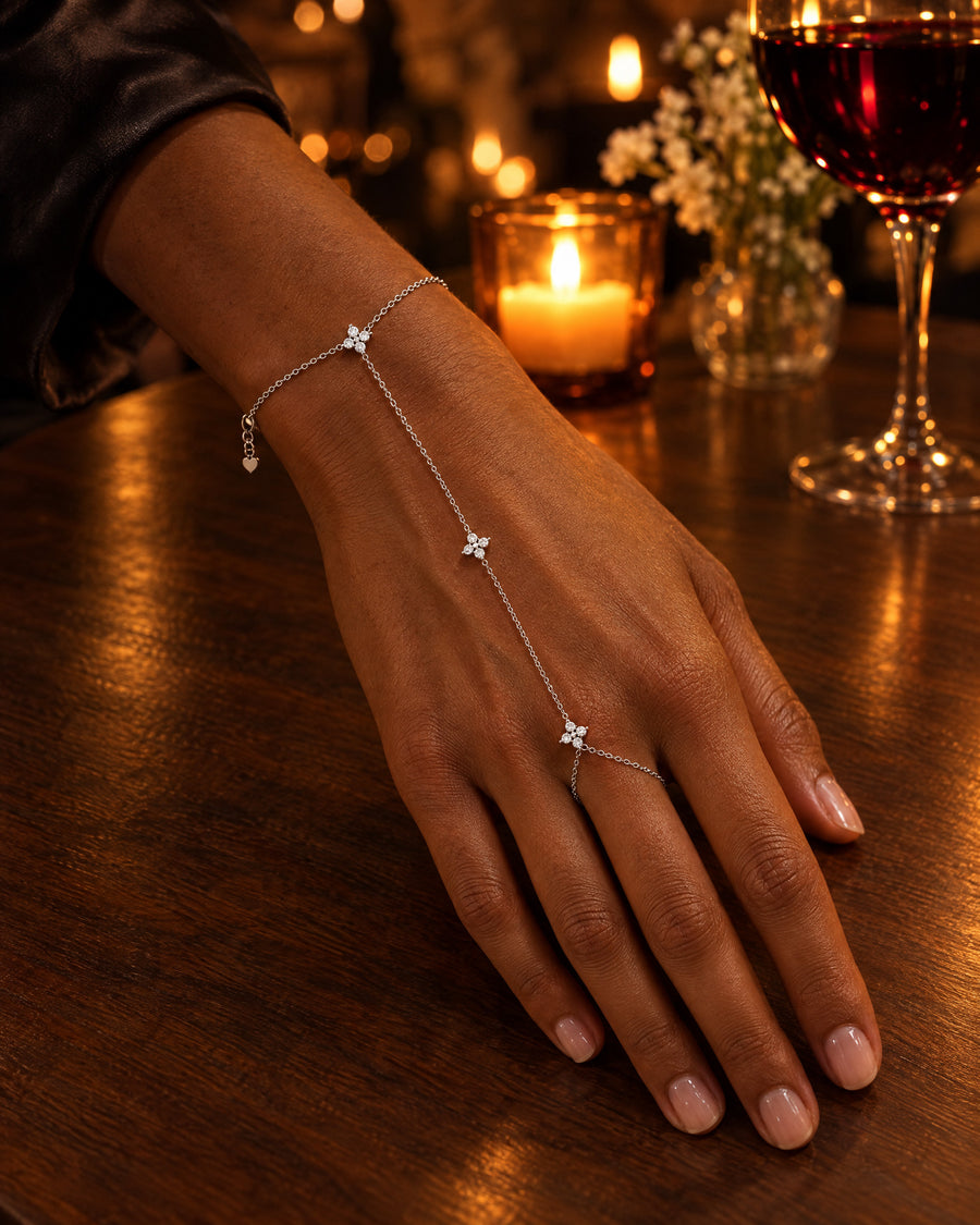 The silver hand chain on a woman's hand resting on a walnut table by candlelight, the three stars at the wrist, the back of the hand and the middle finger, a glass of red wine behind.