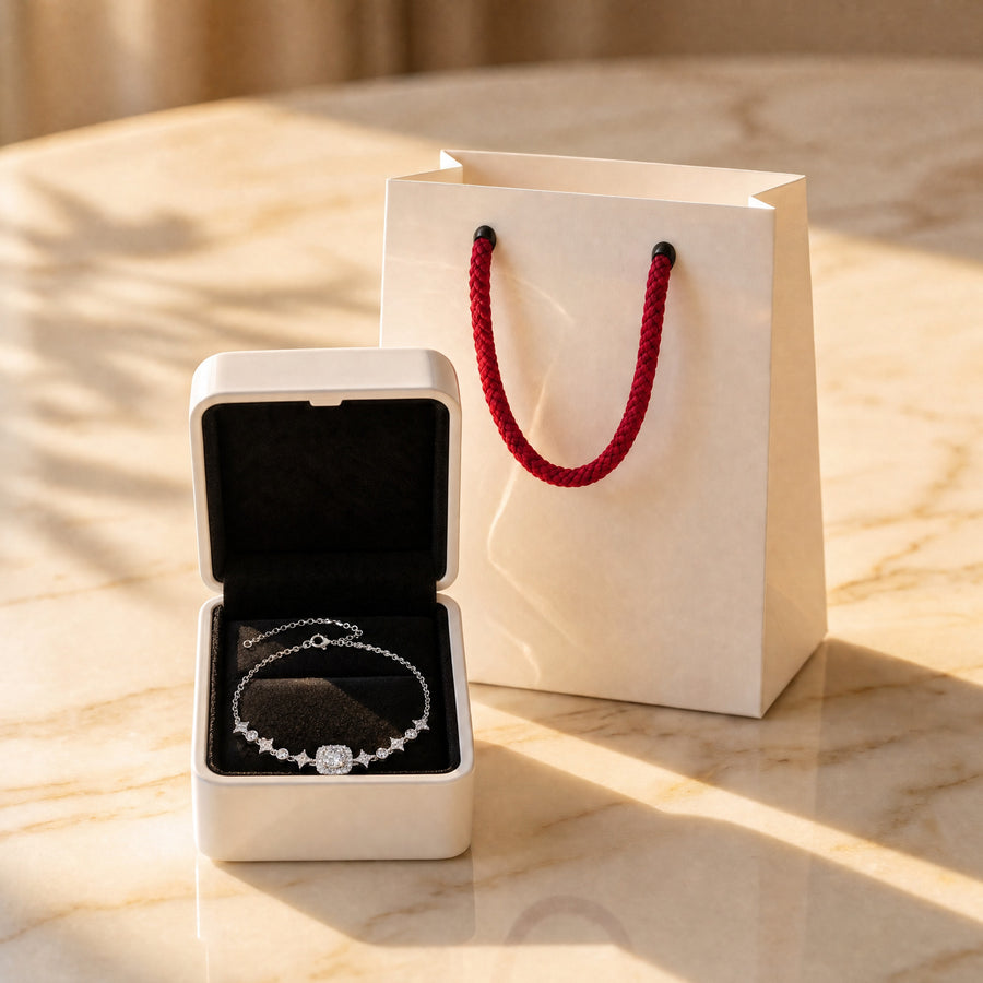 The open white box holding a Starlight bracelet beside the white paper bag with red rope handles on a sunlit marble table.
