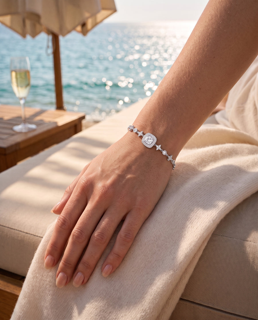 A beach-club daybed in the sun: her hand on a cream cashmere throw, the sea behind.
