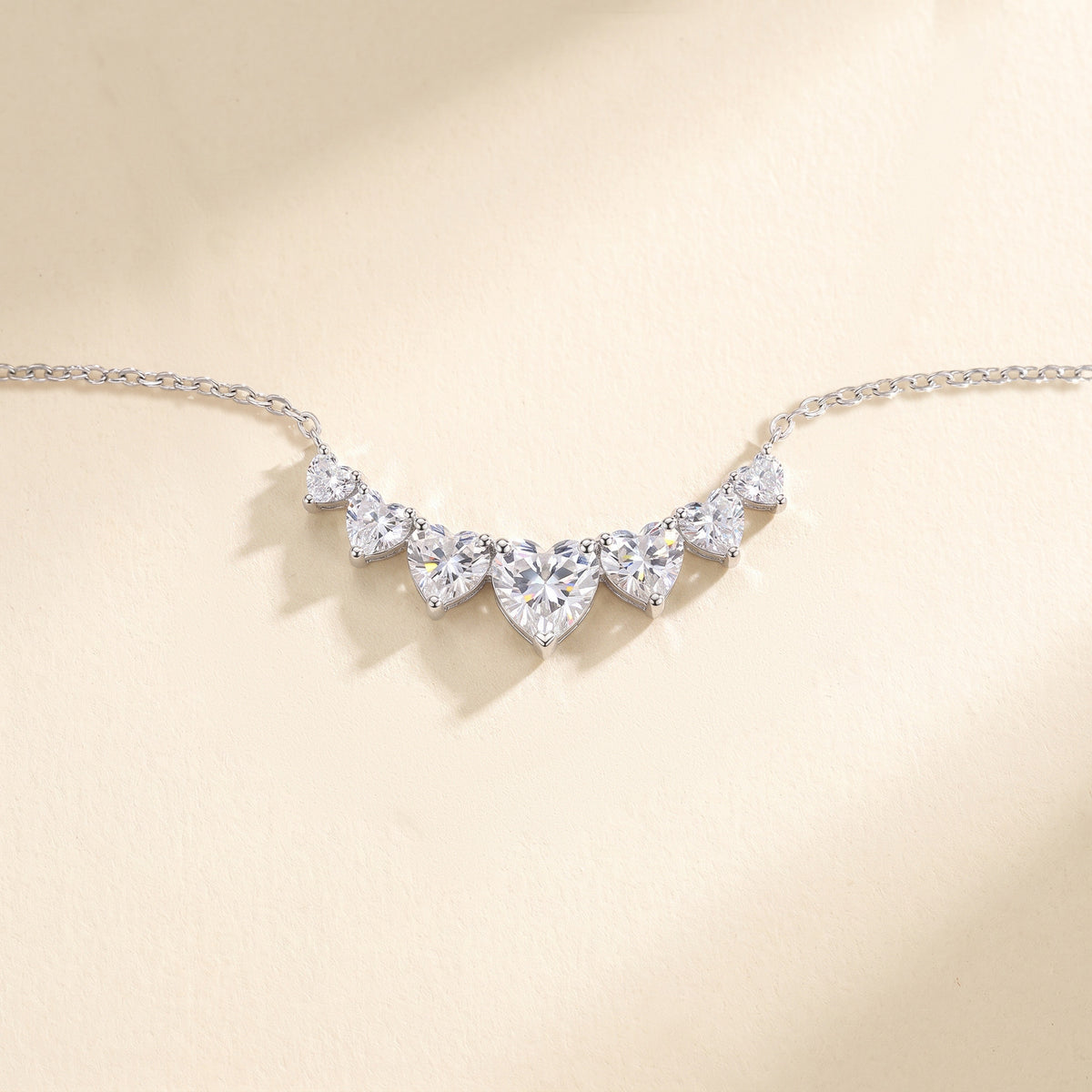 The Mossante Sweetheart Necklace laid on a warm cream ground: a fine silver cable chain running out to both sides from one curved row of seven heart-shaped moissanites, the largest heart at the center and the others stepping down in size to each end, every heart point-down in its own prongs.