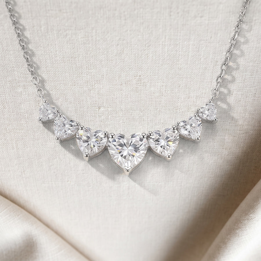 Close view of the seven heart-shaped moissanites in one curved row on pale linen, the largest heart at the center, each in its own silver prongs, the fine chain rising from both ends.