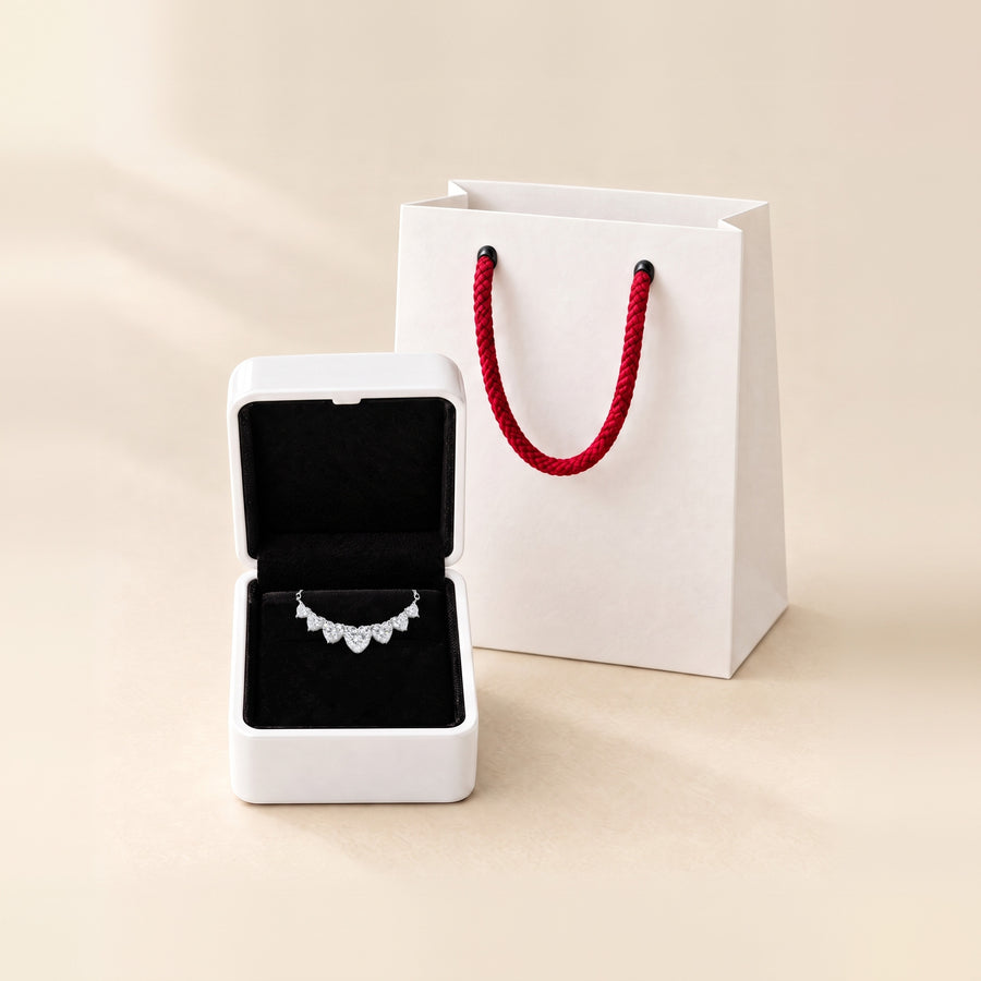 The sweetheart necklace in its open white box, the seven hearts on the black velvet pad and the chain tucked into the slot, the white paper bag with red rope handles behind, on a cream ground.